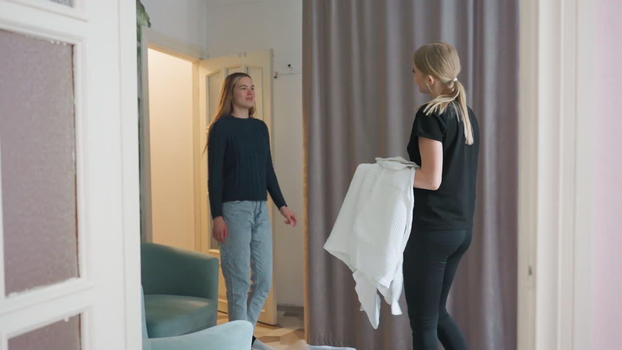 Pretty woman enters warmly lit room with green leafy wallpaper and soft armchairs, greeted by a lady beside the curtain who hands her night robe, capturing calm indoor interaction