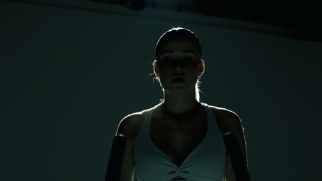 frontal waist-up shot of a woman in a dark studio, lit from behind. the camera pushes in and drops slightly as she ties her hair back, grabs the handlebar, and begins her cycling workout with focus