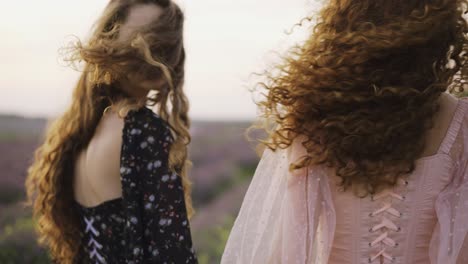 dos mujeres rizadas viajeras están caminando a lo largo de un campo de lavanda en vestidos de corsé