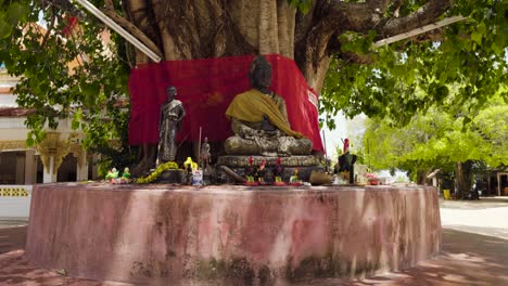 Bodhi-Baum-Mit-Sitzender-Buddha-Statue-Im-Thailändischen-Tempel,-Geschmückt-Mit-Blumen