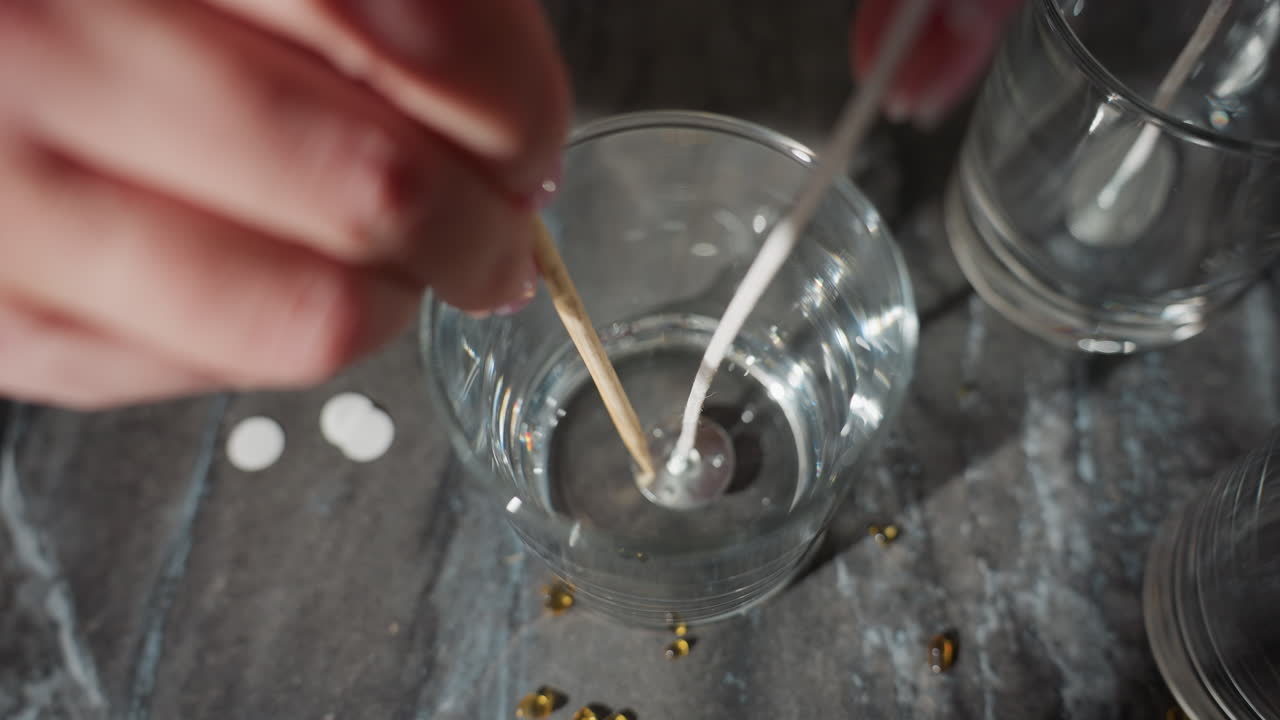 close-up of hands preparing beads design, placing wooden sticks into glass with water, working with beads and design materials, creating delicate craft work on marble countertop