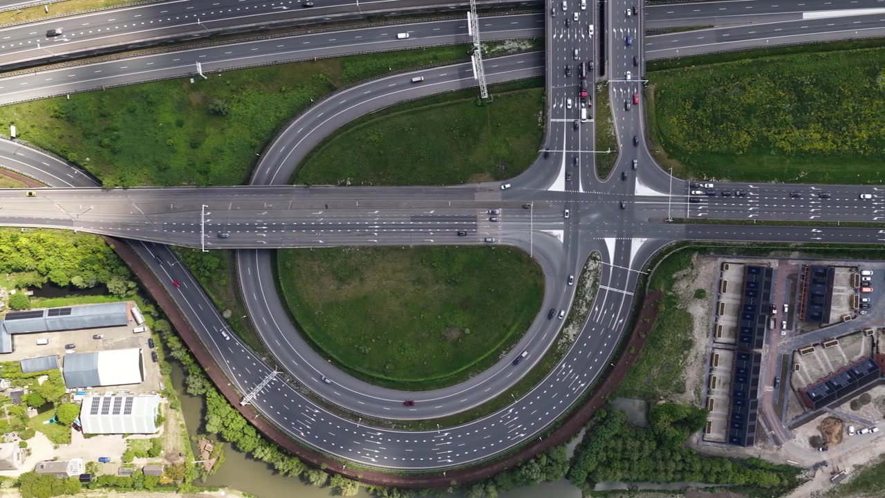 High angle aerial perspective showing a complex road junction with cars and trucks driving along a multi lane highway, overpass, and connecting ramps surrounded by green urban landscape