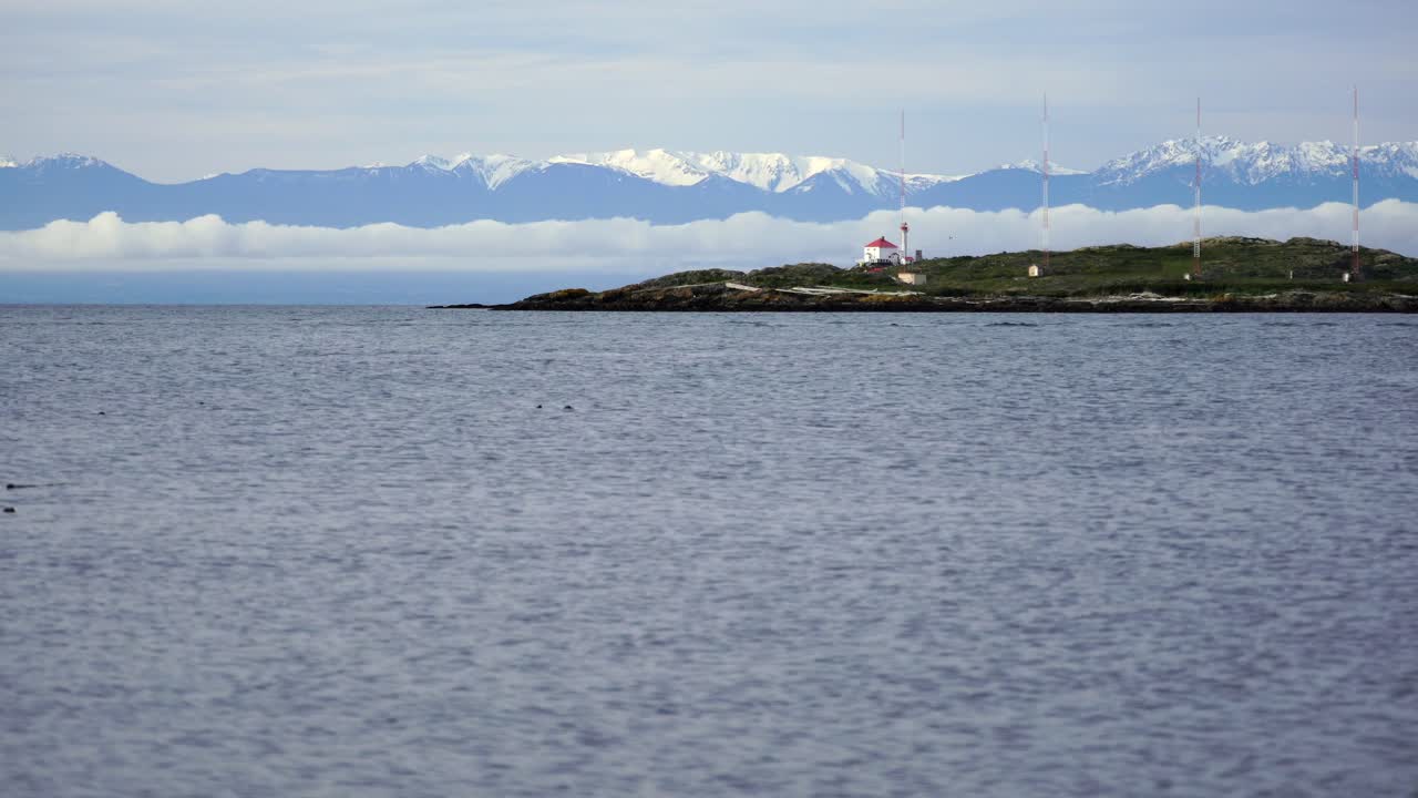 Calm sea with a distant island featuring a lighthouse and antennas. Snow-capped mountains and a boat are visible under a cloudy sky