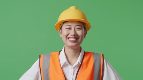 안전 헬을 착용 한 아시아 여성 엔지니어의 클로즈업은 녹색 스크린 배경 스튜디오에서 팔을 들고 서있는 동안 카메라를보고 있습니다.