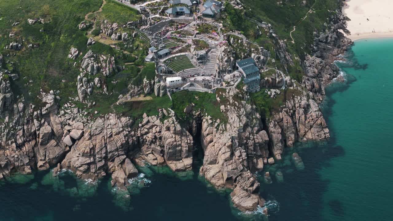 High drone orbit reveals full Minack Theatre carved into Cornwall cliffside with ocean backdrop, aerial establishing overview