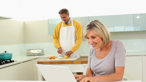 Mujer-Usando-Una-Computadora-Portátil-Mientras-Su-Marido-Corta-Verduras