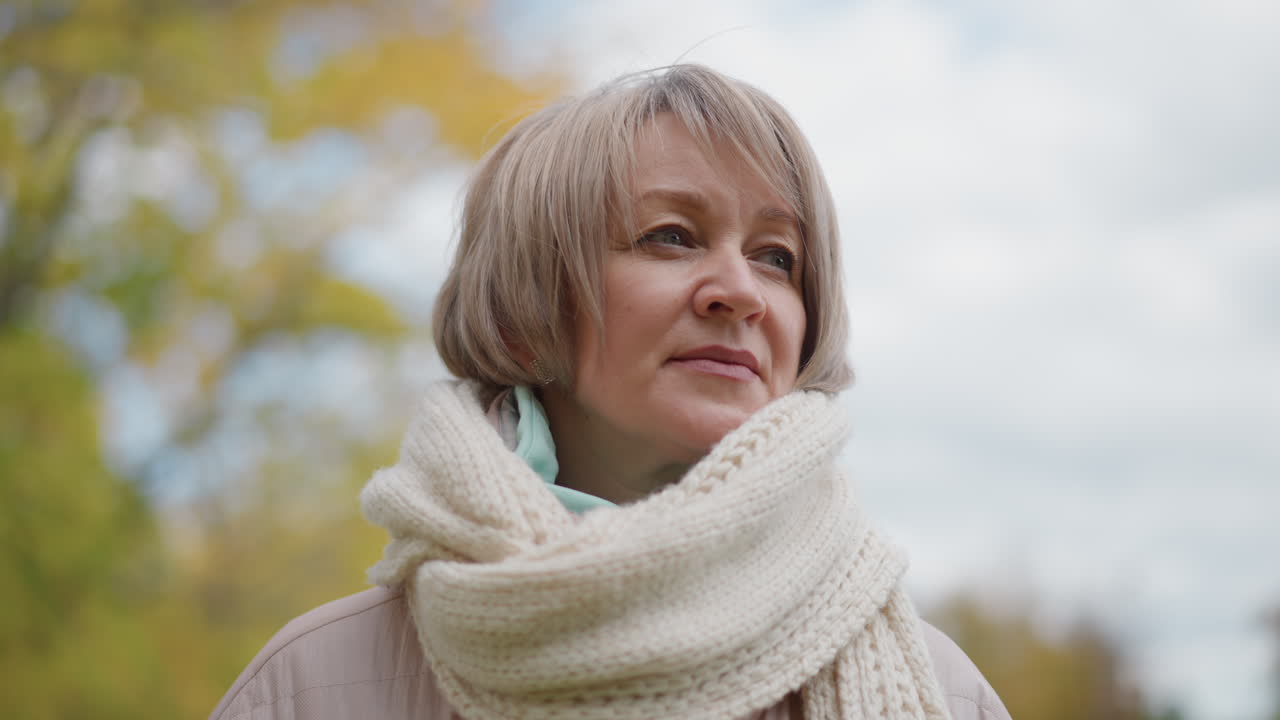 close up of elegant mother looking into distant horizon amidst blurred green foliage and soft autumn sky wearing chunky knit scarf and pastel coat