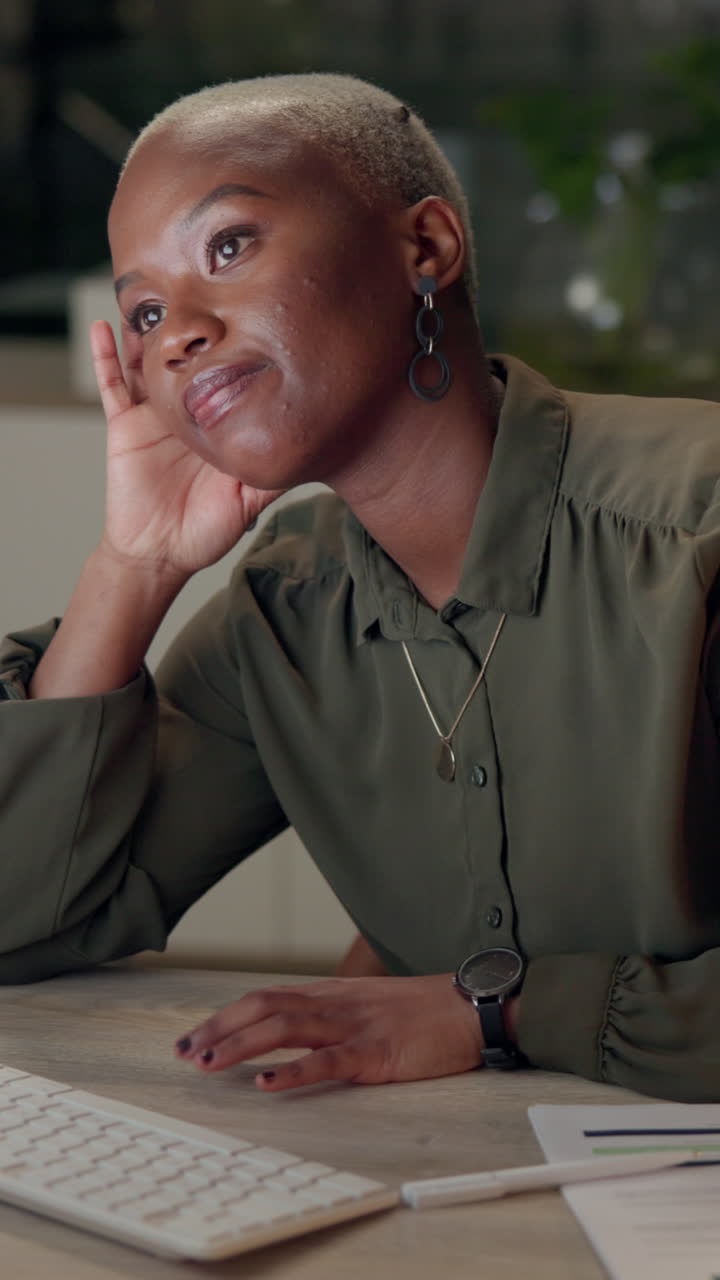 A young woman in a green shirt sits at a desk looking away thoughtfully while working on a computer.