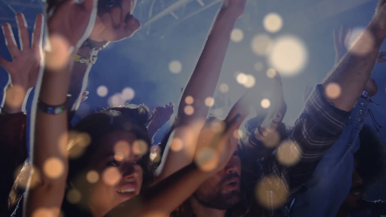 Crowd of concertgoers raising arms as beat dropping, bokeh flares floating across crowd