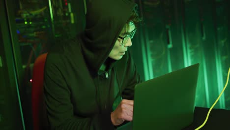 asian male hacker in hoodie using laptop by computer servers