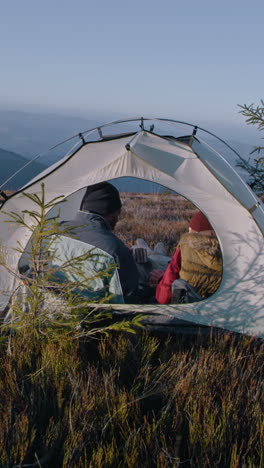 multiethnic tourist couple sit in tent on top of mountain hill: they talk and drink tea. two travelers stopped to rest during adventure vacation. romantic hiker family admire the scenery. vertical shot