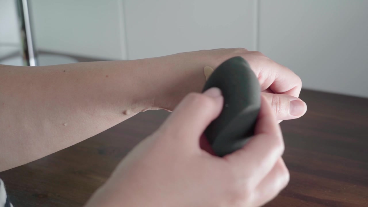 Hands pump liquid foundation onto the back of a hand and tap it with a makeup sponge like a small palette on a wooden table.