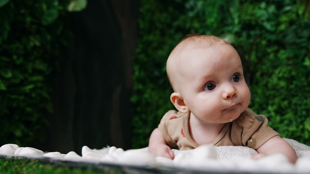 Lovely child lying on the belly looking around with surprise. Adorable Caucasian baby with big eyes. Close up. Greenery at backdrop.