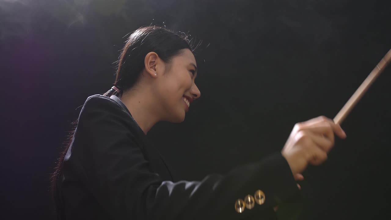 안개가 가득한 검은 스튜디오에서 눈을 감고 몸짓을 하는 아시아계 지휘자 여성의 측면 시각