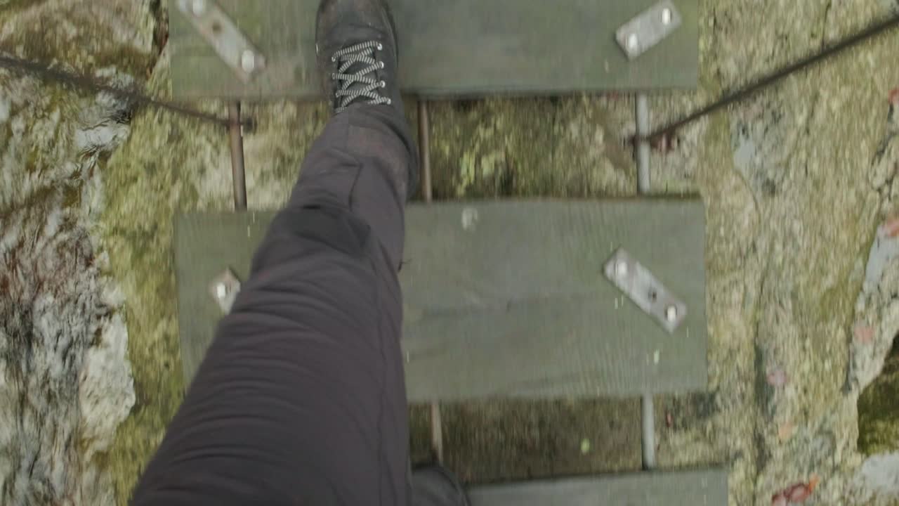 Man walking over a wooden bridge with holes in the wood. Heavy flow of water underneth. Camera looking down to see the shoes walking over the bridge in Slovenia