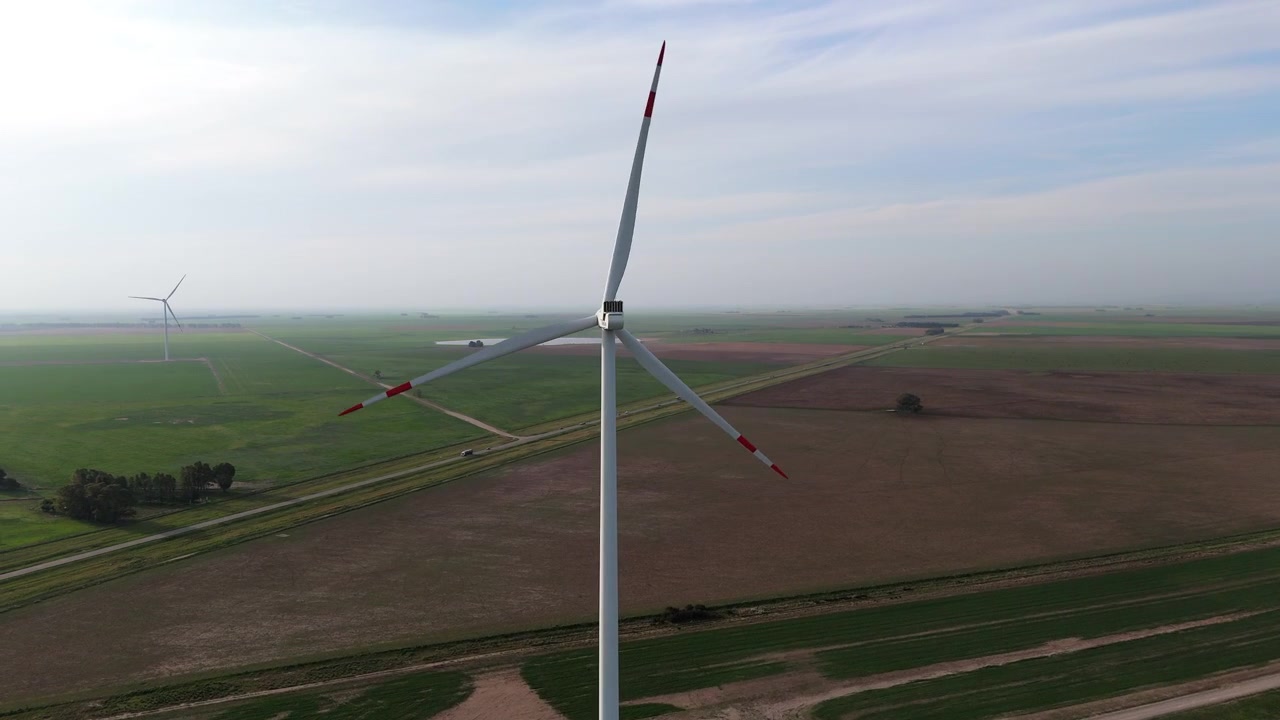 Una singola pala eolica in primo piano, più turbine eoliche sullo sfondo, luce diurna, nebbioso, campi marroni e verdi intorno, spazio per il testo, energia verde.