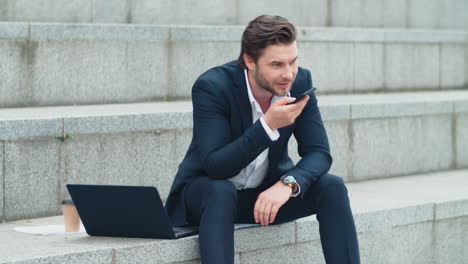 Business-man-recording-audio-message-on-cellphone-outdoors.Man-sitting-on-stairs
