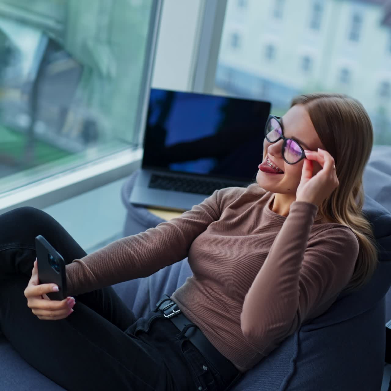 Young lady sitting in bean bag chair fooling around. Woman is taking funny selfie on her phone. High angle view