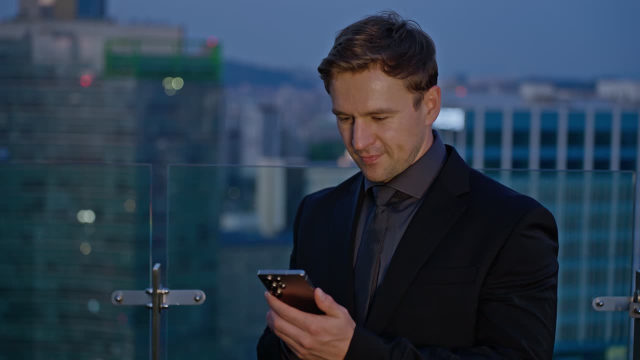 Smiling businessman in black suit using smartphone joyfully on rooftop terrace with modern city skyline at dusk