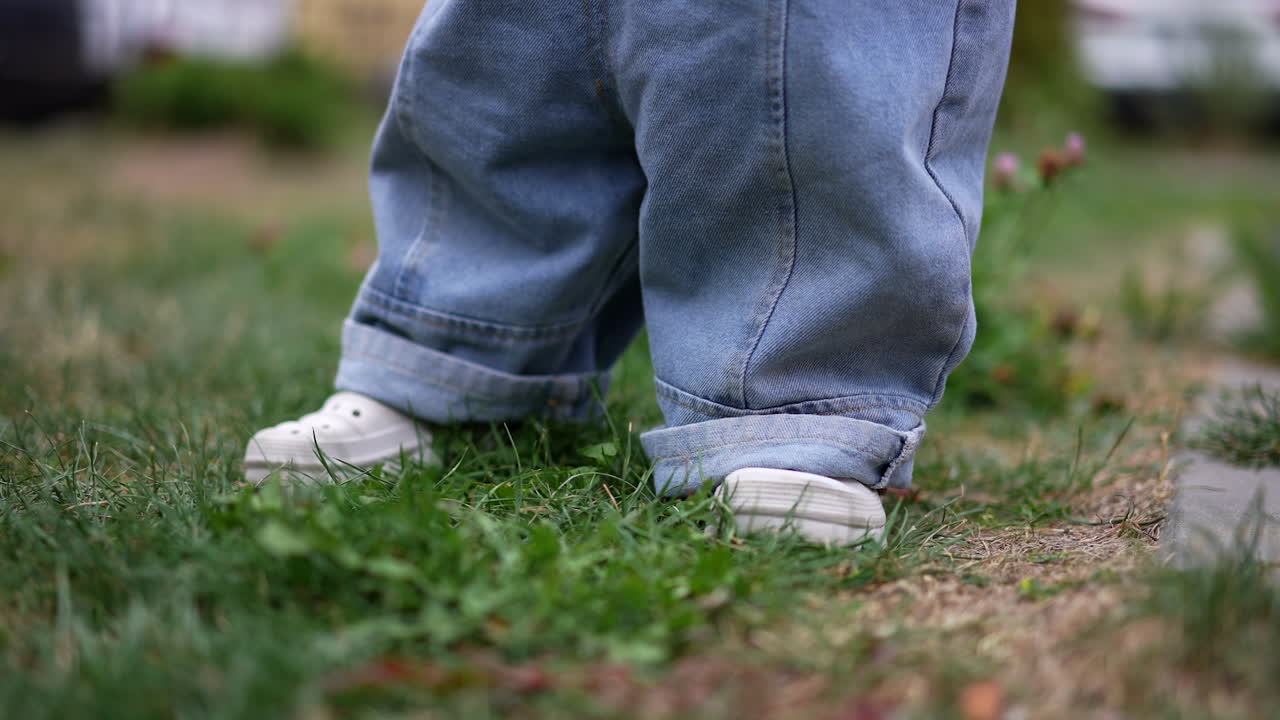 Lovely blond Caucasian toddler in jeans romper sits near the flowers. Baby boy plays outdoors in summer. Blurred backdrop.