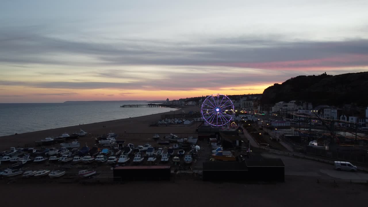 Drone flies toward illuminated Ferris wheel over boats on Eastbourne beach at dusk