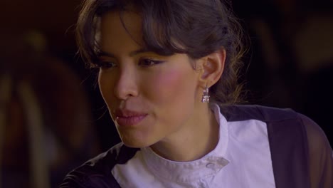 cinematic close up shot of a beautiful woman preparing herself for a horse ride, smiling and wearing a black and white horse riding outfit with a horse in the background, slow motion, slomo