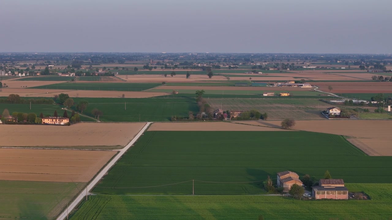 Terreni agricoli coltivati a mosaico si estendono attraverso la Pianura Padana, Italia, con cascine rurali, campi agricoli e strade di campagna che formano un vasto paesaggio produttivo, rivelazione con pullback del drone.