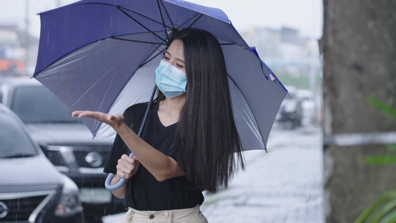 젊은 아시아 여성은 얼굴 마스크를 착용하고 우산을 들고 길 에 서 있습니다. 비가 오는 날, 샤워, 아시아의 비가 오는 계절, 도로 에 주차된 차, 그녀의 팔을 어 비방울을 잡습니다.