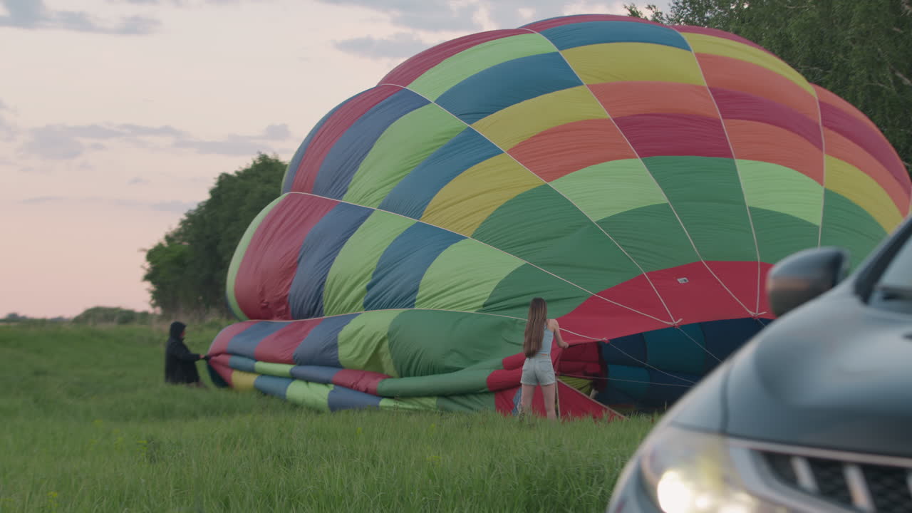 Close up tall grass framing hot air balloon carrying people and pilot heating air, floating low above lush open field under gentle pastel soft calm dawn sky, distant tree line visible