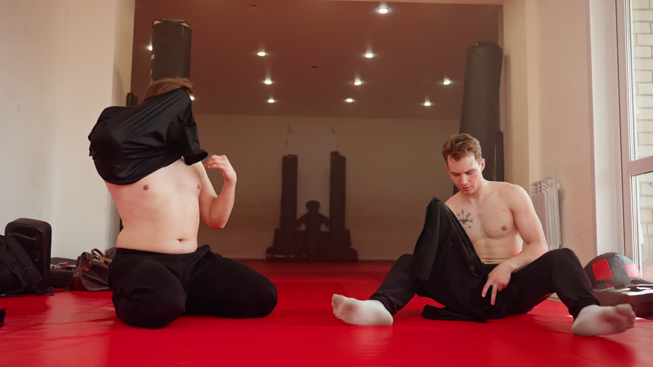 Two men sitting on red mat in gym after training session, wearing their shirts, resting and preparing to change clothes, muscular build, sports bags in background, fitness and athletic lifestyle moment