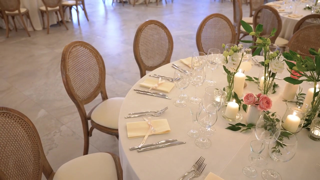 Wide angle view of an elegantly set banquet table for a wedding reception, featuring soft candlelight, pink roses, lush greenery, and sparkling glassware in a bright, airy hall.