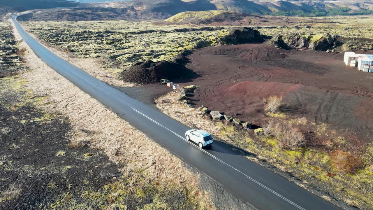 imagen volando sobre islandia, viendo un vehículo atravesar musgo y roca volcánica, con montañas cubiertas de nieve contra un cielo azul claro, evocando el deseo de viajar