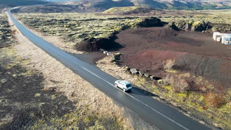 Picture-soaring-over-Iceland,-watching-a-vehicle-traverse-moss-and-volcanic-rock,-with-snow-capped-mountains-against-a-clear-blue-sky,-evoking-wanderlust