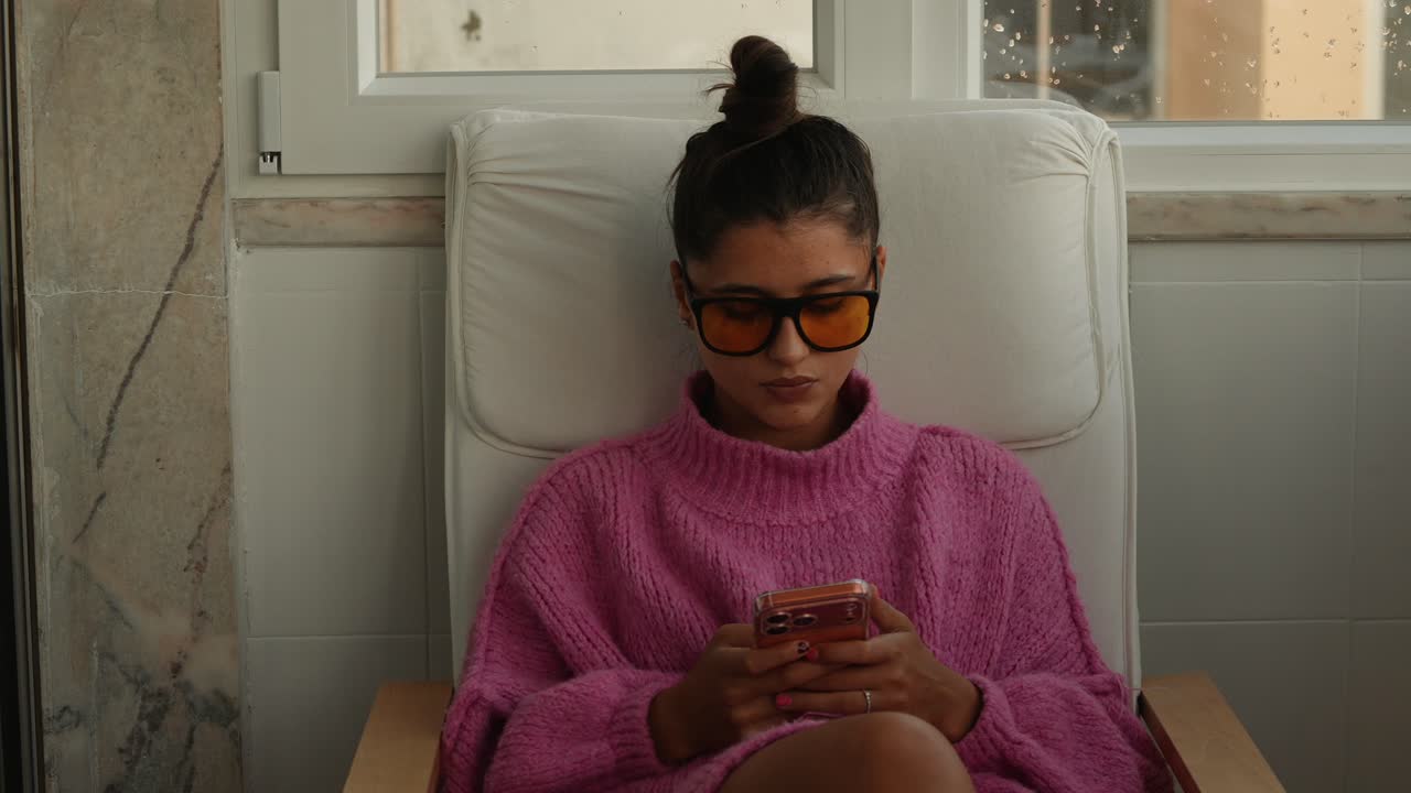 Giovane donna in maglione rosa che usa lo smartphone seduta