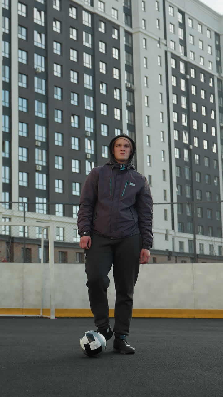 Sportsman performing ankle twists with left and right legs, balancing ball in front on urban sports arena with goalpost and tall urban building in background