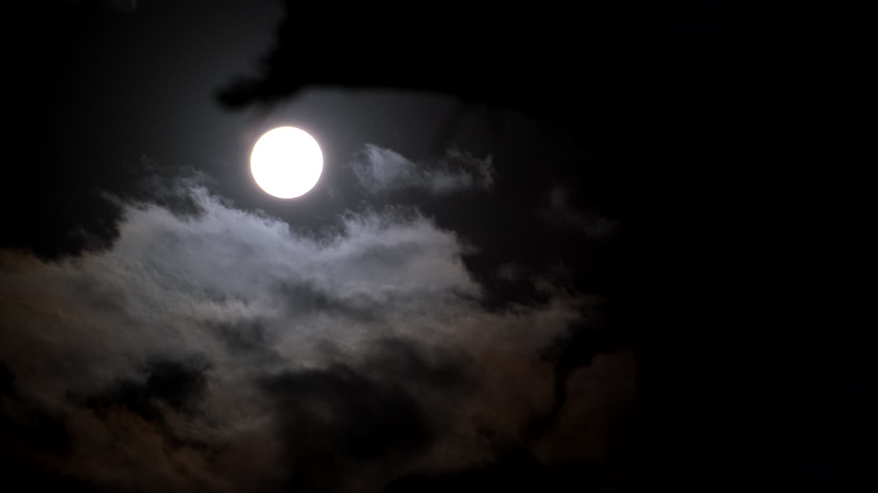 Bright round orb of full moon in dark night sky as clouds ominously pass by