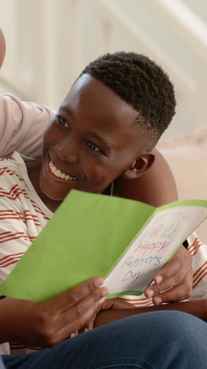 Vertical video: Holding green Father's card, child in striped shirt reading on couch, adult leaning