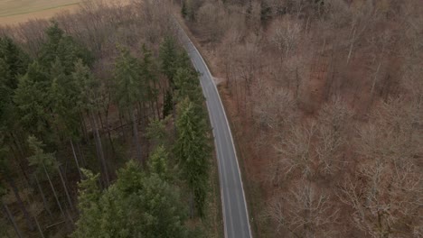 Vista-De-Pájaro-De-Un-Automóvil-Que-Viaja-A-Lo-Largo-De-Una-Carretera-Rural-Curva-A-Través-De-Un-Bosque-Mixto-Y-Parcialmente-Desnudo