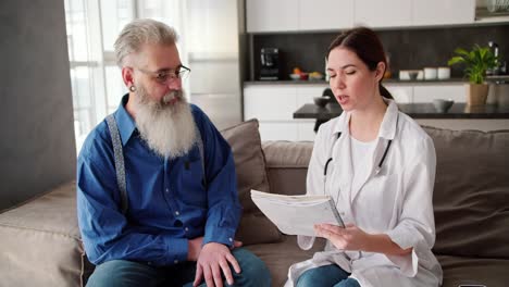 A-man-with-gray-hair-of-advanced-age-with-a-lush-gray-beard-in-a-blue-shirt-sits-on-a-modern-sofa-near-a-brunette-doctor-girl-in-a-white-coat-who-tells-him-about-methods-of-maintaining-health-in-a-modern-apartment