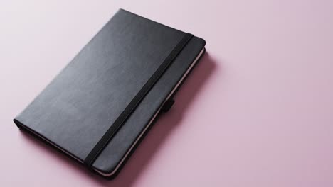 Close-up-of-closed-black-notebook-with-copy-space-on-pink-background-in-slow-motion