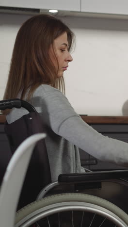 mujer joven seria se sienta en silla de ruedas cerca de la mesa limpieza apartamento. concentrado ama de casa aspiradoras en la cocina mantener el orden en el hogar cuidadosamente vista lateral