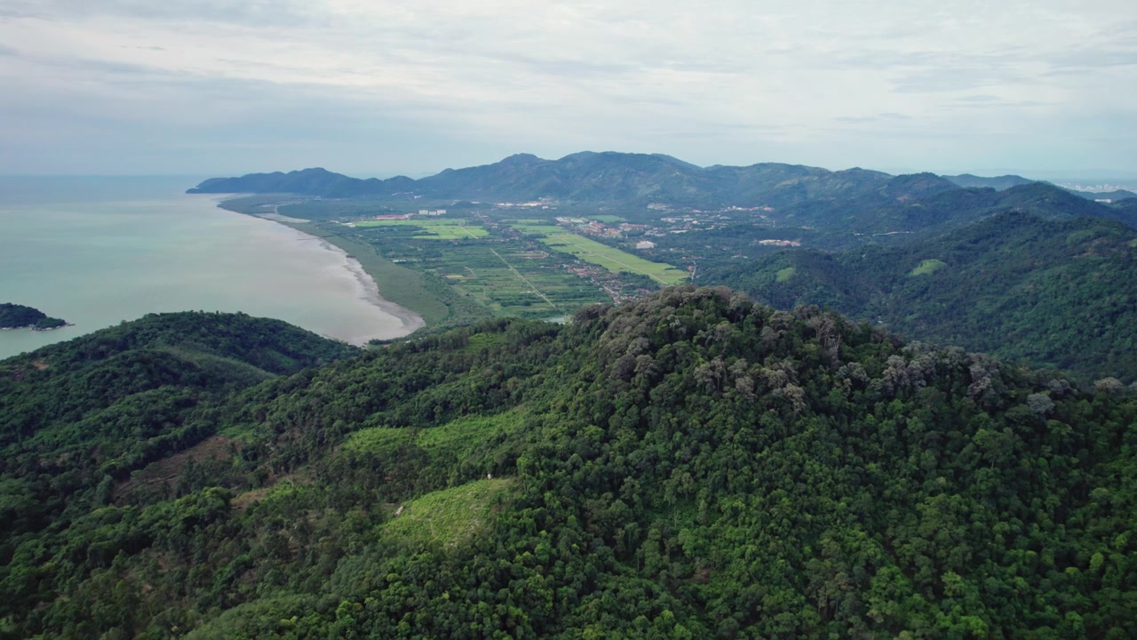 High aerial footage showcases the dramatic landscape of Balik Pulau, with lush mountain ridges overlooking green farmland, rice fields and the western coastline of Penang Island, Malaysia.