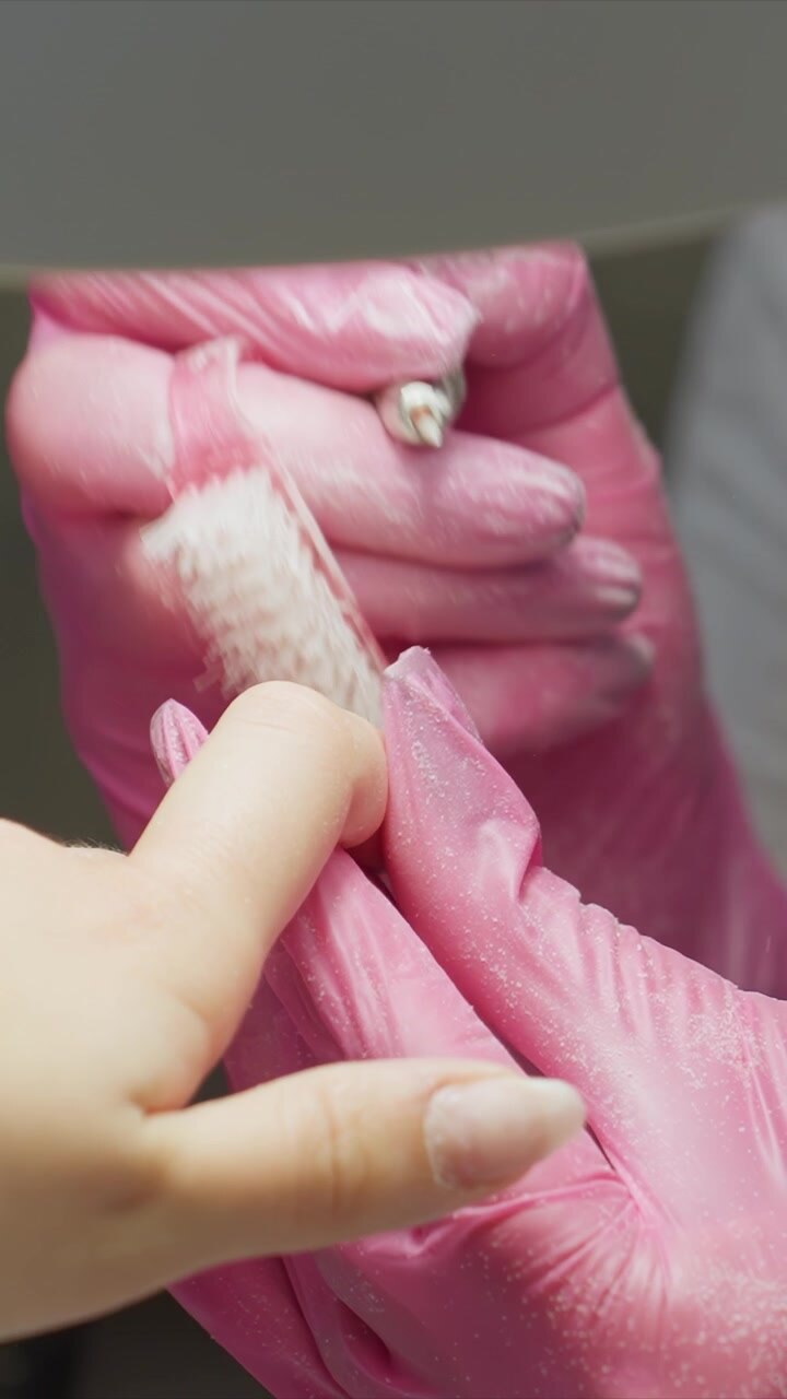 Close up shows person hand being carefully cleaned under pink disposable gloves. Another gloved finger holds nail steady while various cleaning and shaping actions are performed focusing