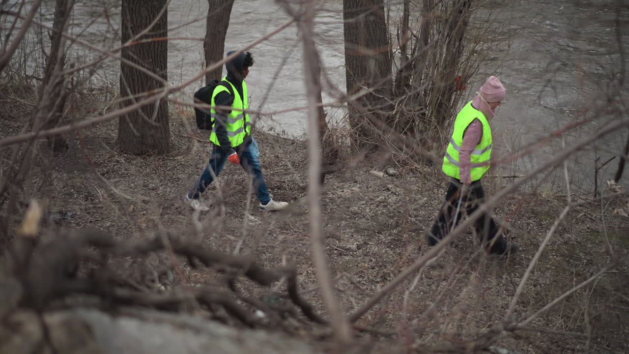 Volunteers wearing bright reflective vests walking along polluted riverbank during environmental cleanup, carrying bags with collected trash and plastic waste, navigating through bare trees on cold