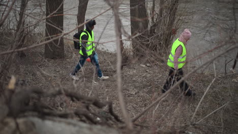 volunteers wearing bright reflective vests walking along polluted riverbank during environmental cleanup, carrying bags with collected trash and plastic waste, navigating through bare trees on cold