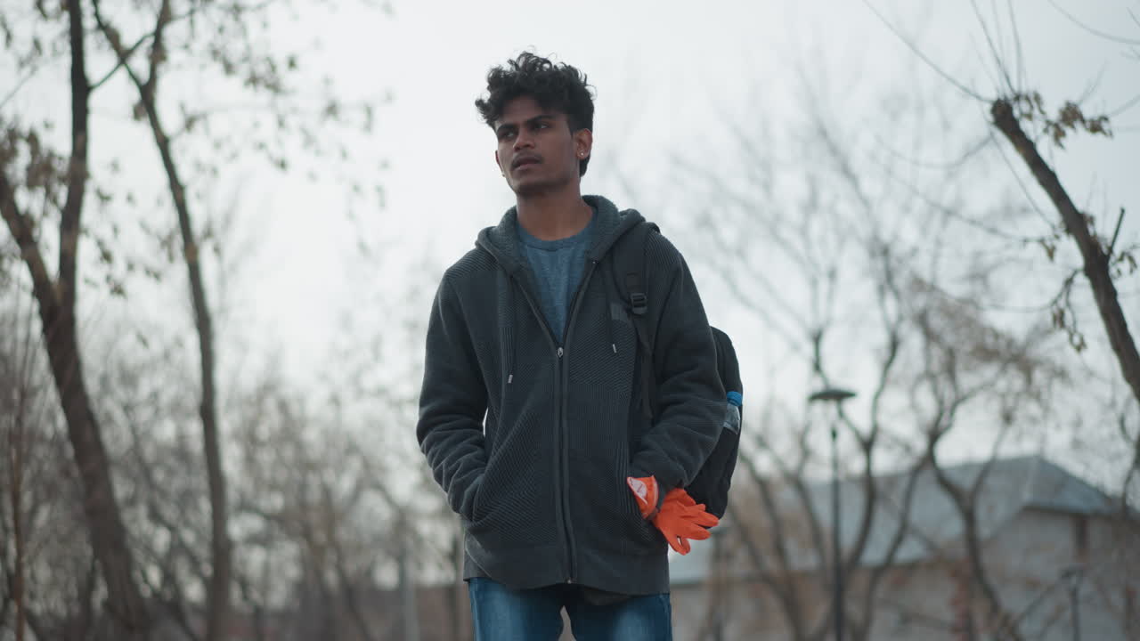 Un giovane uomo in piedi all'aperto in un tranquillo parco urbano, indossa una felpa scura con cappuccio e uno zaino, tiene guanti arancioni, ha uno sguardo serio e contemplativo, con alberi invernali spogli intorno, in un'atmosfera fredda e con le mani in tasca.