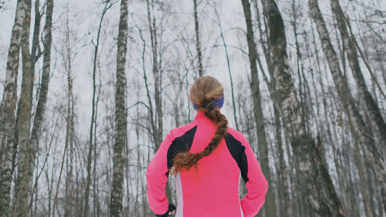 A young woman runs through the park in the winter in a pink jacket. View from the back