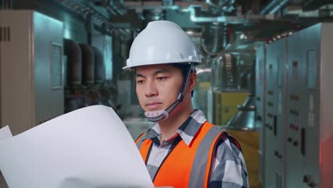 close up side view of asian male engineer with safety helmet looking at blueprint in his hands and looking around while standing in engine control room, work of electrical generators