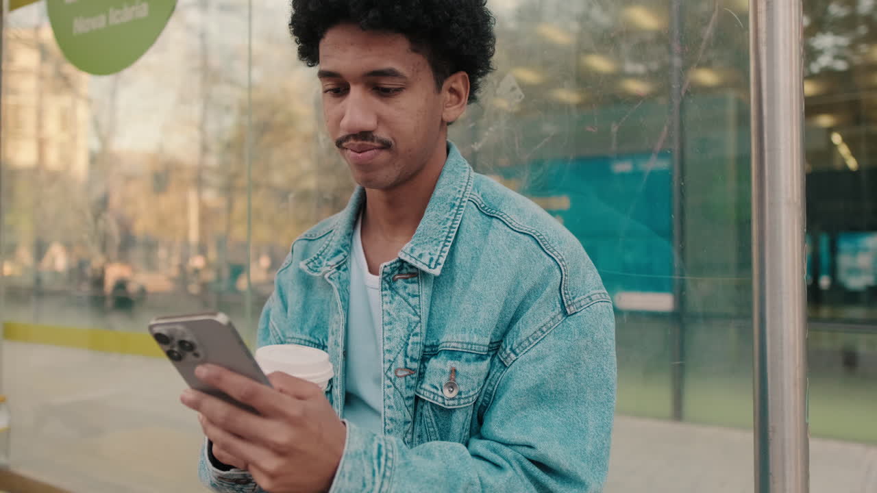 Giovane uomo che usa lo smartphone e beve caffè alla fermata dell'autobus