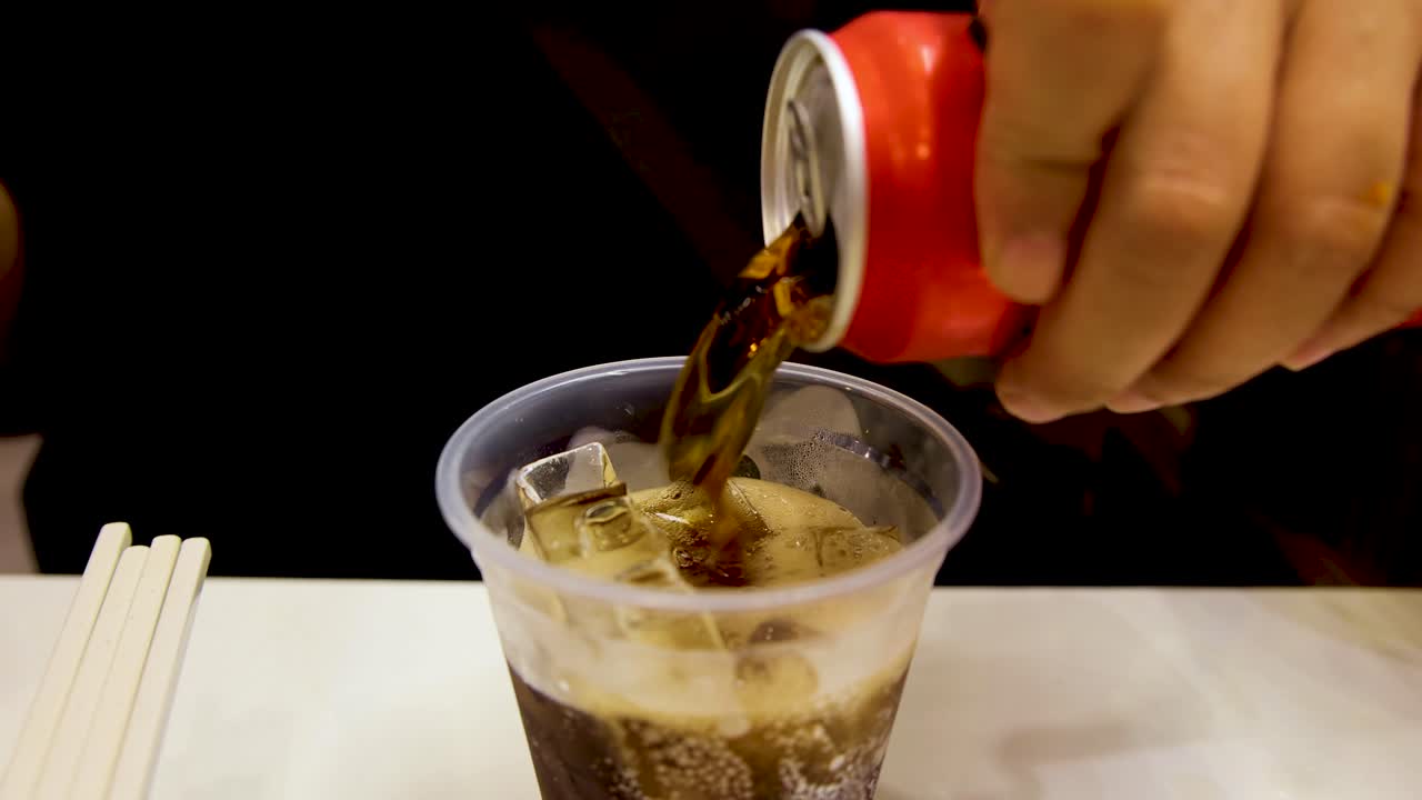 A hand pours dark cola from a red can into a clear plastic cup filled with ice cubes on a white table under bright indoor lighting